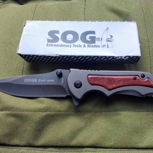 May include: A SOG Flash Tanto folding knife with a black blade and a gray handle with a wood inlay. The knife is next to its box, which has the SOG logo and the text "Extraordinary Tools & Blades."