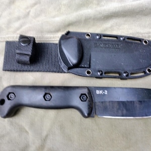 May include: A black knife with a sheath and a black nylon strap. The knife has a black handle and a blade. The sheath is black and has the text "KA-BAR" on it. The knife is labeled "BK-2".