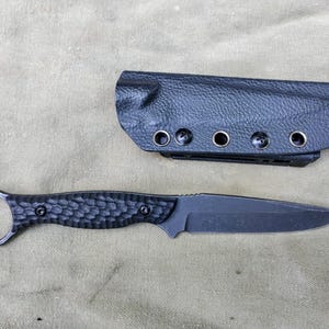 May include: A black fixed-blade knife with a textured handle and a finger ring, accompanied by a black sheath. The knife has a sharp blade and is designed for utility purposes. The sheath has multiple eyelets for mounting.