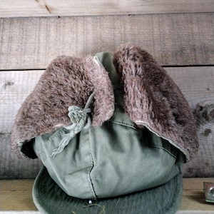 May include: An olive green winter hat with ear flaps lined with brown faux fur. The hat has a rounded crown and a brim. A drawstring is visible at the front. The hat is set against a wooden background.
