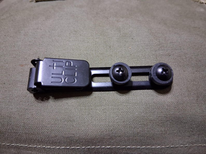 May include: A black metal clip with the word "ULTI CLIP" embossed on the top. The clip has two screws on a bar. The background is a textured, olive-green fabric.