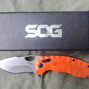 May include: An orange and silver folding knife with a partially serrated blade, next to a black box with the white letters "SOG". The knife has a textured orange handle and a silver blade. The box is rectangular and has a textured surface.