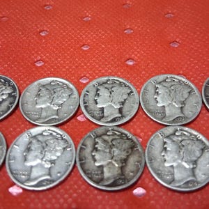 May include: Ten silver Mercury dimes arranged on a red surface. Each coin features a profile of a woman wearing a winged cap. The coins are slightly tarnished, showing signs of age and use.