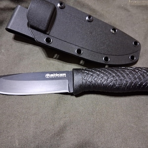 May include: A black Boker Solingen knife with a textured handle and a matching sheath. The blade has a silver edge and the Boker logo is visible. The knife and sheath are on a dark green surface.