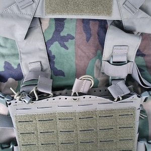 May include: Close-up of a tactical vest in a camouflage pattern. The vest features multiple straps, buckles, and hook-and-loop patches in shades of gray and olive green. The background displays a woodland camouflage fabric.