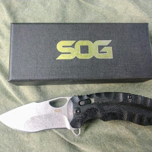 May include: A black and silver folding knife with a textured handle and a silver blade. The knife is next to a black box with the gold letters "SOG" on it. The knife is approximately 7 inches long.
