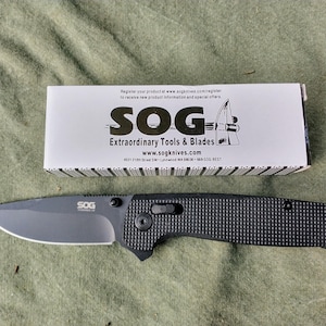 May include: A black SOG folding knife with a textured handle and a gray blade lies next to its product box. The box is white with black text and the SOG logo. The knife is a tactical tool.