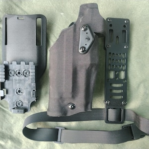 May include: A black tactical holster set, including a holster, belt loop, and mounting hardware. The set features a dark gray holster with a belt and various mounting components. The components are made of durable materials.