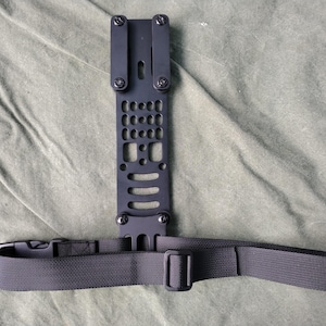 May include: Black metal plate with a strap attached. The plate has a grid pattern and multiple holes. The strap is black nylon with a buckle.