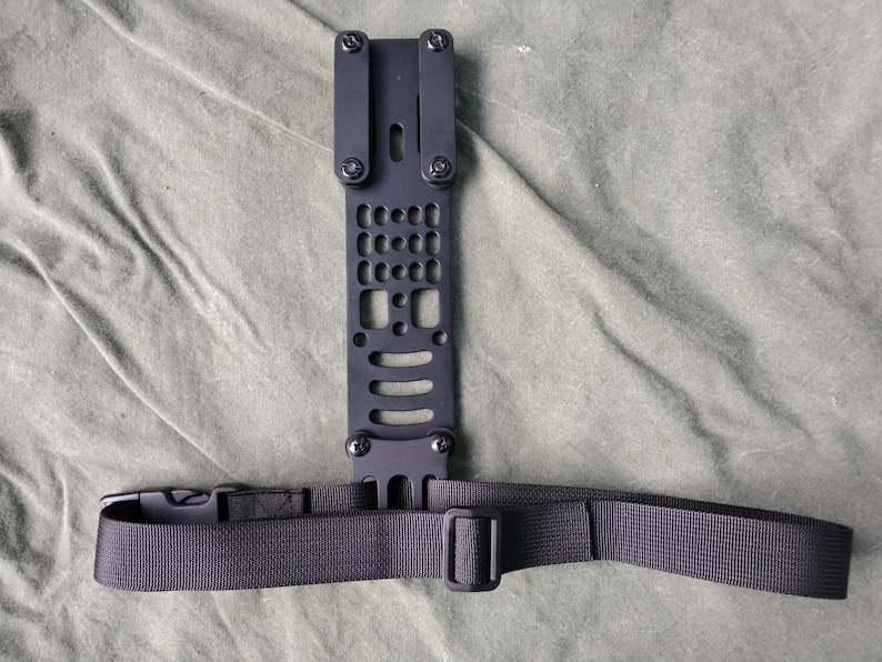 May include: Black metal plate with a strap attached. The plate has a grid pattern and multiple holes. The strap is black nylon with a buckle.