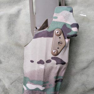 May include: A tactical holster in a camouflage pattern, featuring shades of green, brown, and tan. The holster has a rigid, olive-drab colored belt attachment and is secured with metal hardware. The design suggests a military or outdoor gear theme.