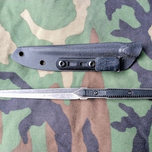 May include: A silver and black dagger with a black sheath. The dagger has a pointed blade and a textured black handle. The sheath is black and has a clip. The background is camouflage.
