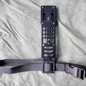 May include: A black metal plate with multiple holes and a black nylon strap with a buckle. The plate has a rectangular shape and is attached to the strap.