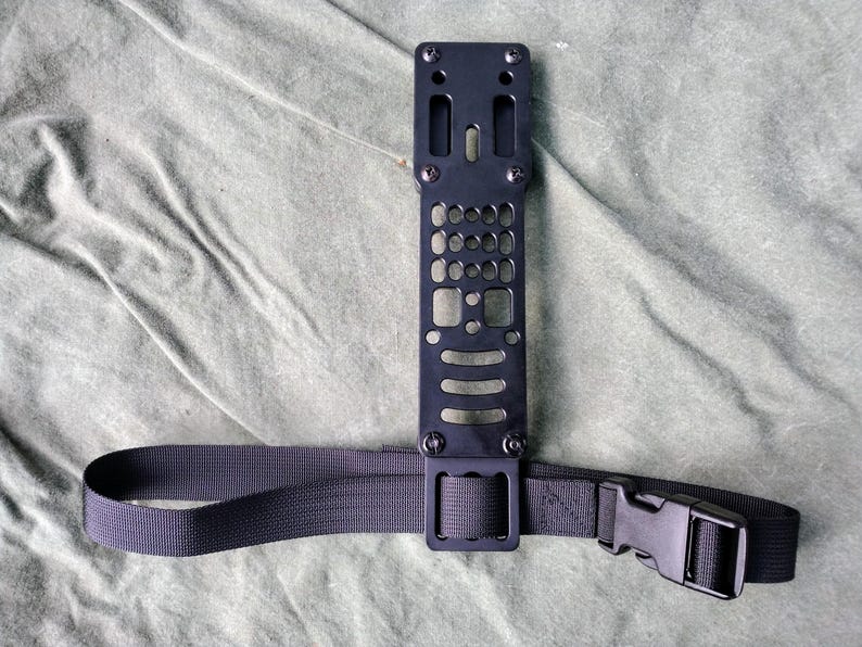 May include: A black metal plate with multiple holes and a black nylon strap with a buckle. The plate has a rectangular shape and is attached to the strap.