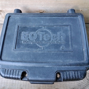 May include: A black, rectangular case with rounded corners and a hinged lid. The case features the text "EOtech Holographic Weapon Sights" in white lettering. The case has a textured surface and two small, cylindrical protrusions on the top.