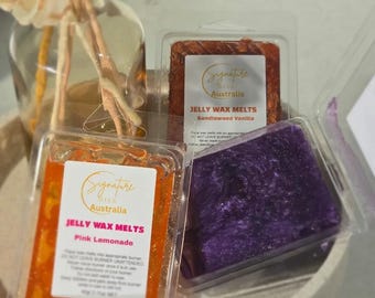Op de afbeelding: Drie doorzichtige plastic bakjes gevuld met jelly wax melts. Eén oranje met het label Pink Lemonade, één bruin met Sandlewood Vanilla en één paars. Elk label bevat de tekst Signature Oils Australia.