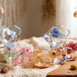 May include: A collection of clear glass mugs with silver handles, each adorned with colorful floral designs. The flowers include blue, pink, orange, and red. The mugs are arranged on a wooden surface, creating a visually appealing display.