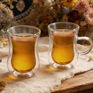 Könnte beinhalten: Zwei transparente Glasbecher, gefüllt mit bernsteinfarbener Flüssigkeit, vermutlich Tee oder Kaffee, stehen auf einer Holzoberfläche. Ein Becher hat einen Henkel. Die doppelwandigen Becher stehen vor getrockneten Blumen und gemustertem Stoff.