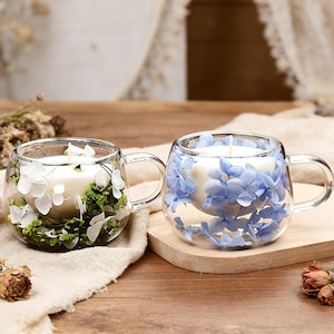 Op de afbeelding: Twee heldere glazen mokken met handvatten, elk met een witte kaars. Eén mok is versierd met witte bloemen en groen blad, de andere met blauwe bloemen. De mokken staan op een houten oppervlak.