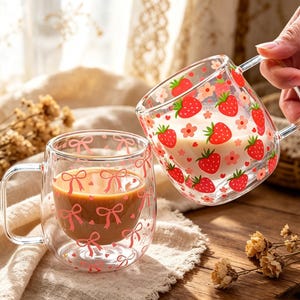 Könnte beinhalten: Zwei durchsichtige doppelwandige Glastassen. Eine Tasse ist mit einem Muster aus roten Erdbeeren und kleinen rosa Blumen verziert und mit weißer Flüssigkeit gefüllt. Die andere Tasse zeigt rosa Schleifen und kleine Herzen mit brauner Flüssigkeit.
