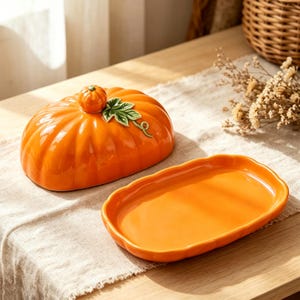 May include: A glossy orange ceramic butter dish with a lid shaped like a pumpkin. The lid features a small pumpkin stem handle and a sculpted green leaf detail. The base is an oval-shaped orange tray with a smooth, slightly scalloped edge.