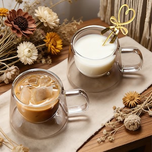 May include: Two clear double-walled glass mugs, one filled with iced coffee and the other with milk, sit on a beige cloth. A gold heart-shaped spoon rests in the milk. Dried flowers and a wooden surface complete the scene.