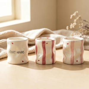 Könnte beinhalten: Drei Keramikbecher mit welliger, unebener Oberfläche. Der linke Becher ist cremefarben mit dem Schriftzug "COZY HOURS" und kleinen roten Herzen. Die mittleren und rechten Becher haben vertikale rosa Streifen und kleine rote Herzen auf Creme.