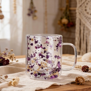 Peut inclure: Une tasse en verre transparent à double paroi, remplie de délicates fleurs séchées violettes et blanches. La tasse a une poignée incurvée et est posée sur une surface de couleur claire, créant une esthétique visuellement attrayante.