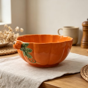 Op de afbeelding: Een glanzende oranje keramische kom in de vorm van een pompoen met een geschulpte rand. De buitenkant is voorzien van een kleine gebeeldhouwde oranje pompoen en een groen blad met een rankdetail aan de zijkant, lijkend op een echte pompoen.