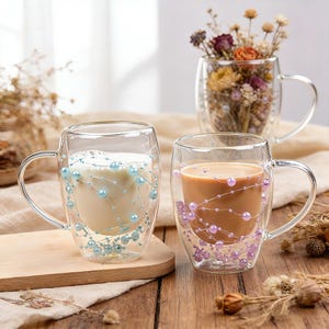 May include: Clear glass mugs with double-walled construction. One mug contains a milky liquid and is decorated with blue beads. Another mug holds a brown liquid and is adorned with purple beads. A third mug holds dried flowers.