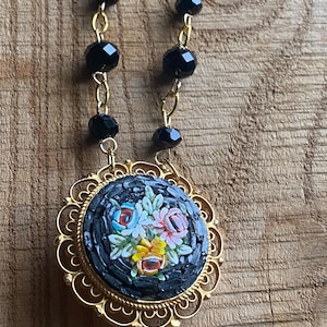 Puede incluir: Un collar dorado con un colgante de mosaico con un diseño floral. El colgante tiene un fondo negro con flores coloridas. La cadena del collar incluye cuentas negras y eslabones dorados.