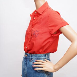 Op de afbeelding: Een rood, kortgemouwd overhemd met knopen en blauwe bloemenborduursel. Het shirt wordt gedragen met een blauwe spijkerbroek.