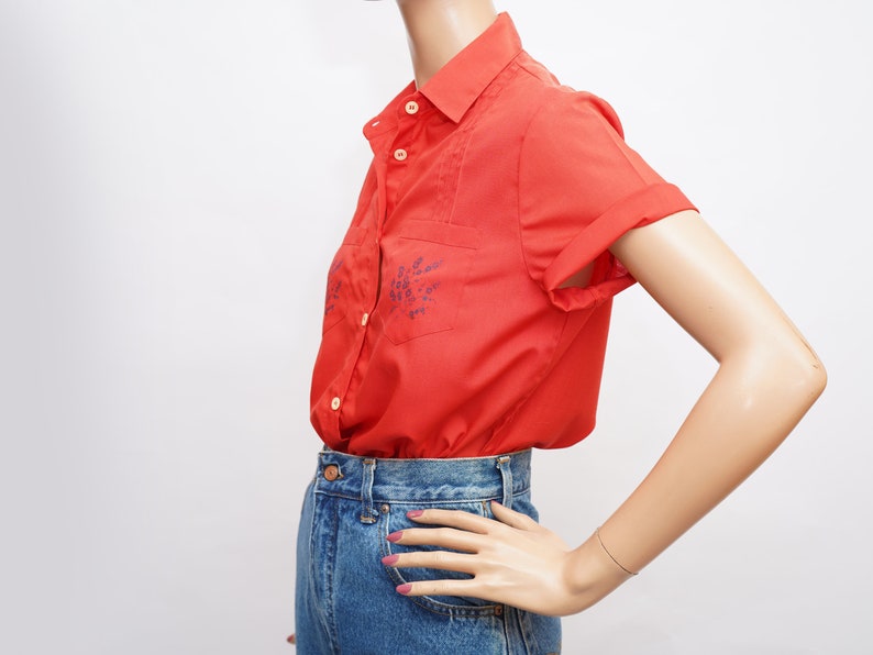 Op de afbeelding: Een rood, kortgemouwd overhemd met knopen en blauwe bloemenborduursel. Het shirt wordt gedragen met een blauwe spijkerbroek.