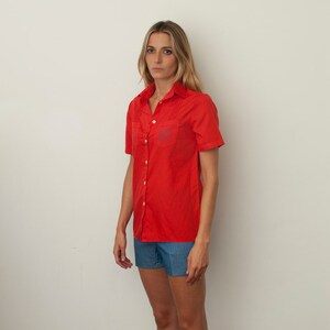 Op de afbeelding: Een vrouw draagt een rood, kort, knoopshirt met een geborduurd blauw logo en een blauwe denim short.