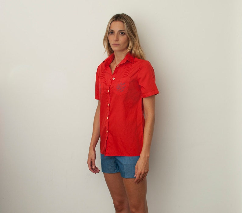 Op de afbeelding: Een vrouw draagt een rood, kort, knoopshirt met een geborduurd blauw logo en een blauwe denim short.
