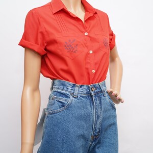 Op de afbeelding: Een rood, kortgeknipt overhemd met knopen en blauwe bloemenborduursel op de voorzakken. Het overhemd wordt gedragen met een hoge blauwe jeans.