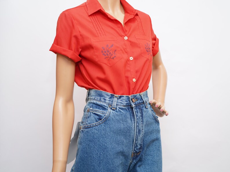 Op de afbeelding: Een rood, kortgeknipt overhemd met knopen en blauwe bloemenborduursel op de voorzakken. Het overhemd wordt gedragen met een hoge blauwe jeans.