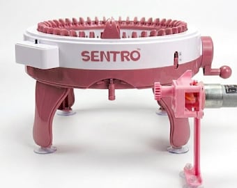 May include: A circular knitting machine with a white and pink plastic frame, featuring the text SENTRO printed on the side. The device includes a hand crank, a series of upright needles along the top rim, and an attached motor component.