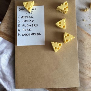 May include: A brown paper shopping list with the words "Shopping List" written at the top. The list includes the following items: apples, bread, flowers, pork, and cucumbers. The list is held in place by five yellow cheese wedge magnets.