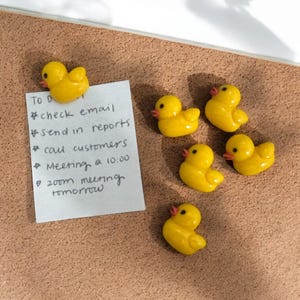 Puede incluir: Pines de corcho de pato de goma amarillo en un tablero de corcho con una lista de tareas escritas a mano. La lista incluye revisar el correo electrónico, enviar informes, llamar a los clientes, reunión a las 10:00, reunión de zoom mañana.