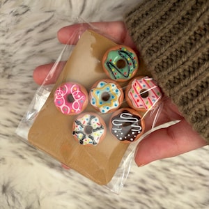 Puede incluir: Una colección de mini donas coloridas en una bolsa de plástico transparente. Cada dona tiene un diseño diferente de glaseado y cobertura, incluyendo rosa, azul, verde y chocolate. Las donas se exhiben sobre un pequeño respaldo de cartón marrón.