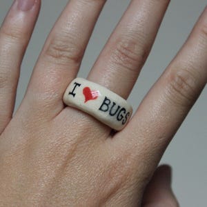 May include: A cream-colored ring with the words "I ❤️ BUGS" in black lettering and a red heart. The ring is worn on a finger.