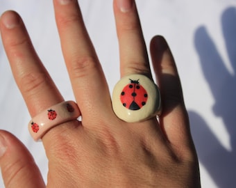 May include: Two rings with ladybug designs. One ring is a light pink band with multiple ladybug illustrations. The other ring is a cream-colored, round ring with a single ladybug illustration in red and black.