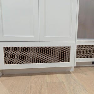 Könnte beinhalten: Weiße Küchenschränke mit dekorativen, geometrisch gemusterten Lüftungsabdeckungen. Die Schränke haben ein modernes Design mit hellem Holzboden. Die Lüftungsabdeckungen sind in einem kontrastierenden Braunton gehalten.