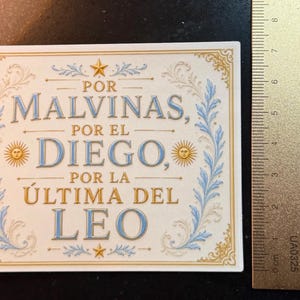 May include: A rectangular sticker with a white background, gold and light blue decorative borders, and the text "POR MALVINAS, POR EL DIEGO, POR LA ÚLTIMA DEL LEO" in gold and blue. A metal ruler shows the sticker is approximately 3.5 inches wide.