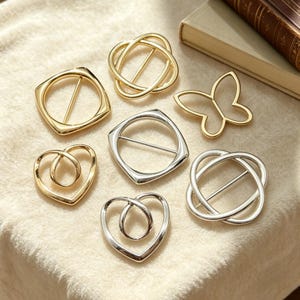 May include: An assortment of decorative metal buckles in gold and silver tones. The buckles feature various designs, including squares, hearts, butterflies, and intertwined shapes. The buckles are displayed on a soft, neutral-colored surface.