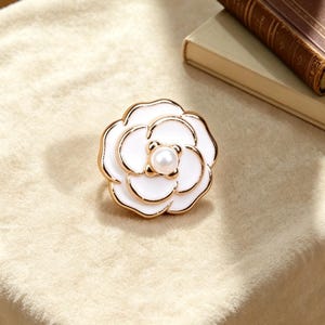 Puede incluir: Broche blanco en forma de flor con ribete dorado y perla central. La broche está sobre una superficie beige, con un libro en el fondo. El diseño floral es un accesorio clásico.
