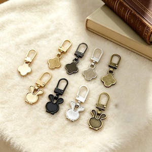 May include: A collection of metal zipper pulls in various colors, including gold, silver, black, and bronze. The pulls feature clover and bunny designs. The pulls are displayed on a soft, white surface.