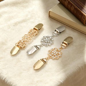 Peut inclure: Trois pinces à pull décoratives en or et argent. L'une présente un motif ajouré, tandis que les autres ont des ornements floraux avec des strass transparents. Les pinces sont conçues pour attacher les vêtements.