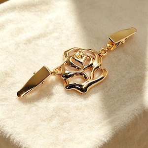 May include: A gold-colored metal scarf clip featuring a rose design. The clip has a detailed rose in the center and two rectangular clasps on either end. The clip is shown on a cream-colored fabric.